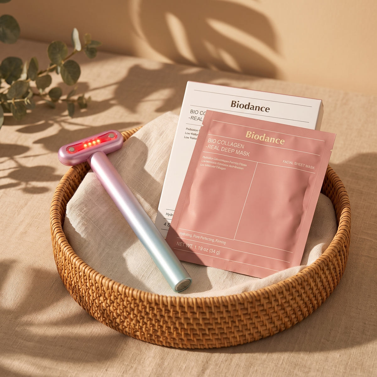 Varita facial Aurora con luz roja junto a la caja y un sobre de mascarilla Biodance en una cesta de ratán